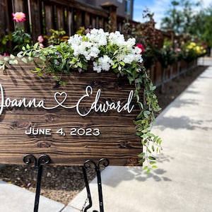 May include: A wooden wedding sign with the names "Joanna & Edward" and the date "June 4, 2023" in white script. The sign is decorated with white flowers and greenery and is displayed on a black metal stand.