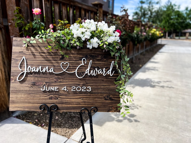 May include: A wooden wedding sign with the names "Joanna & Edward" and the date "June 4, 2023" in white script. The sign is decorated with white flowers and greenery and is displayed on a black metal stand.