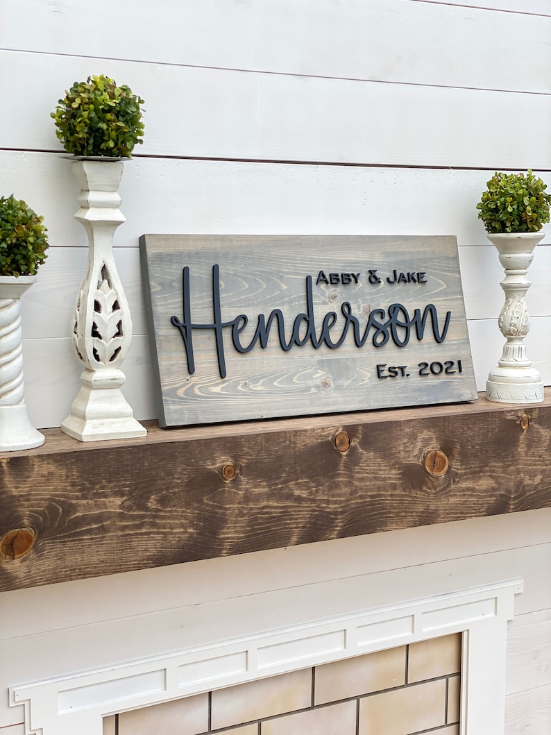 May include: A gray wooden sign with the text "Abby & Jake Henderson Est. 2021" in black raised lettering.