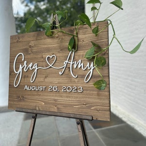 May include: A wooden wedding sign with the names "Greg & Amy" in white script, a heart symbol, and the date "August 26, 2023." The sign is displayed on a wooden easel and adorned with green leafy vines.