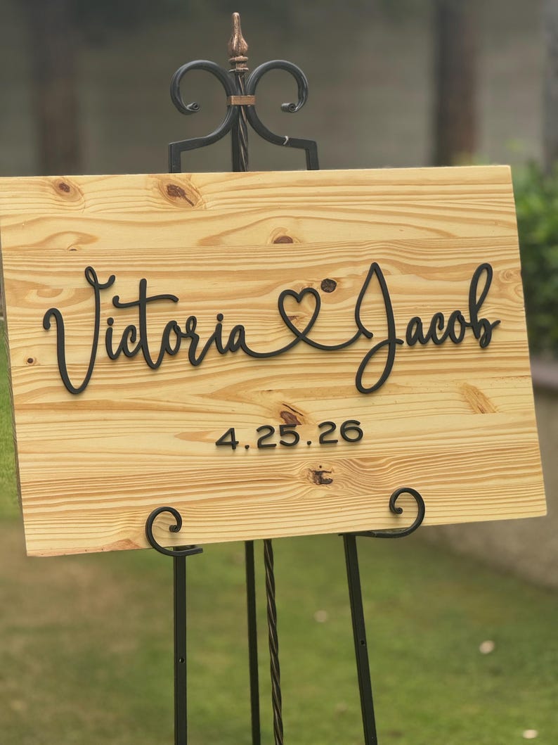 May include: A wooden wedding sign with the names "Victoria & Jacob" in a cursive font, connected by a heart symbol. The date "4.25.26" is below the names. The sign is displayed on a black metal easel.