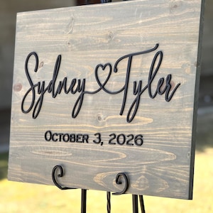 May include: A wooden wedding sign with the names "Sydney & Tyler" in black script, with a heart symbol between the names. The date "October 3, 2026" is displayed below. The sign is mounted on a black metal easel.