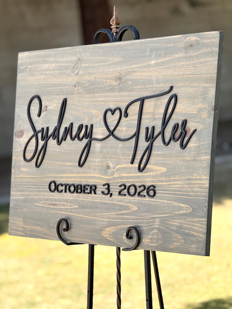 May include: A wooden wedding sign with the names "Sydney & Tyler" in black script, with a heart symbol between the names. The date "October 3, 2026" is displayed below. The sign is mounted on a black metal easel.