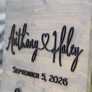 May include: A wooden sign with the names "Anthony & Haley" in black script, joined by a heart symbol. Below, the date "SEPTEMBER 5, 2026" is displayed in a bold, sans-serif font. The sign has a gray-toned finish.