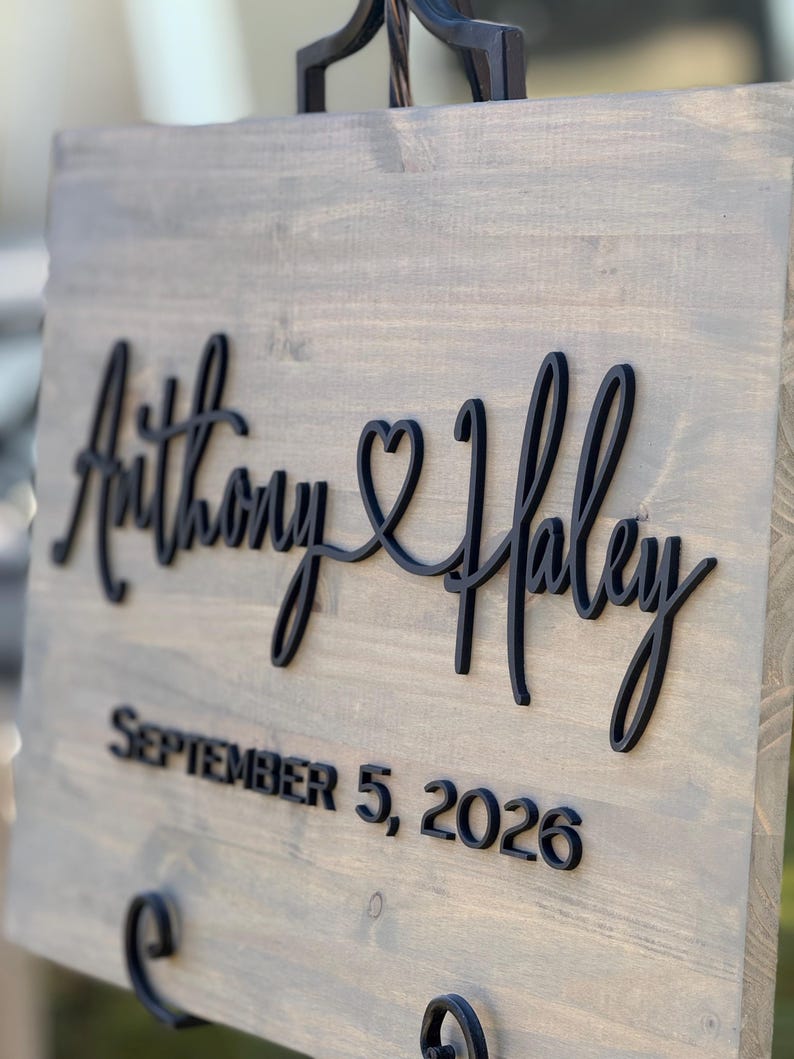 May include: A wooden sign with the names "Anthony & Haley" in black script, joined by a heart symbol. Below, the date "SEPTEMBER 5, 2026" is displayed in a bold, sans-serif font. The sign has a gray-toned finish.