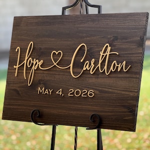May include: A dark brown wooden sign with the names "Hope & Carlton" in gold script, with a heart symbol between the names. The date "MAY 4, 2026" is below the names. The sign is displayed on a black metal easel.