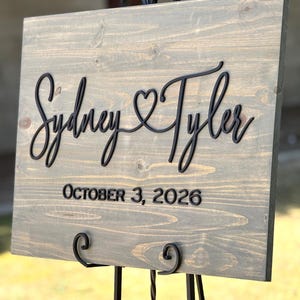 May include: A wooden wedding sign with the names "Sydney & Tyler" in a cursive font, with a heart symbol between the names. The date "October 3, 2026" is below the names. The sign is displayed on a black metal easel.