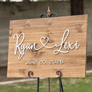 May include: A wooden wedding sign with the names "Ryan & Lexi" in white cursive script, with a heart symbol between the names. The date "JUNE 20, 2026" is printed below. The sign is mounted on a black metal stand.