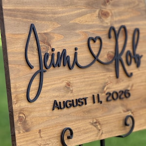 May include: Wooden wedding sign with the names "Jimi & Rob" in a black script font, connected by a heart symbol. The date "August 11, 2026" is displayed below in a sans-serif font. The sign is mounted on a black metal easel.