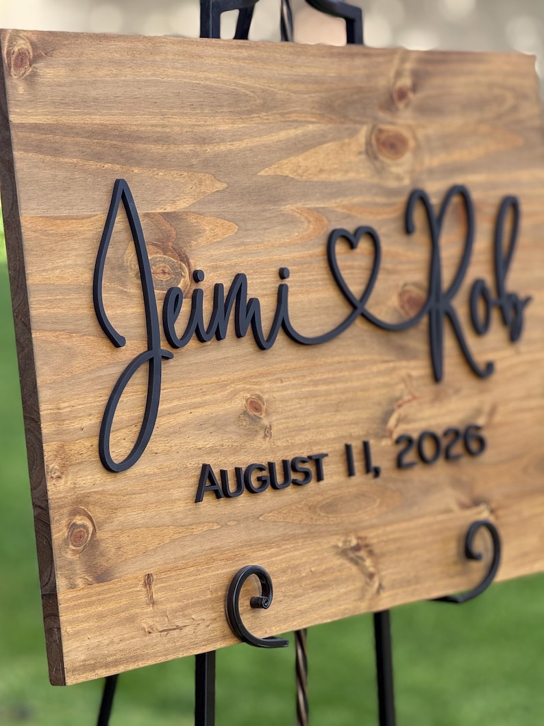 May include: Wooden wedding sign with the names "Jimi & Rob" in a black script font, connected by a heart symbol. The date "August 11, 2026" is displayed below in a sans-serif font. The sign is mounted on a black metal easel.