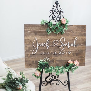 Walnut stained wooden board with white 3D letters with couples’s first names connected with a heart and wedding date below.