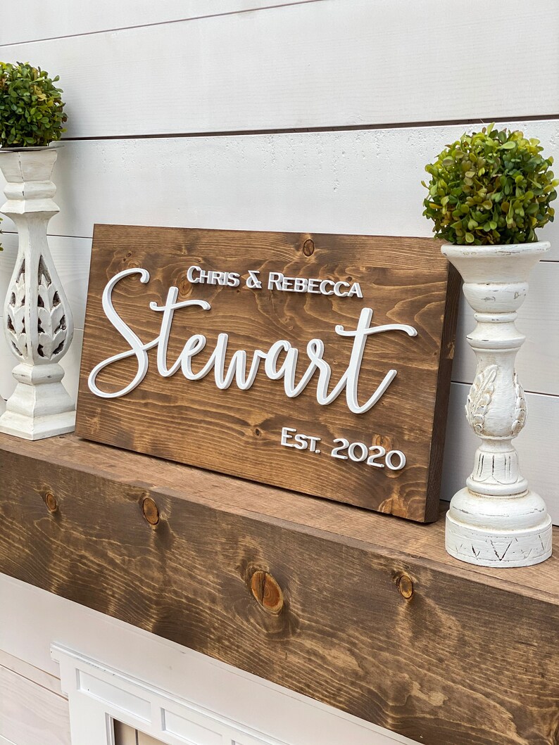May include: A wooden sign with the text "Chris & Rebecca Stewart Est. 2020" in white raised letters. The sign is brown and has a rustic look.