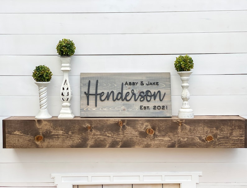 May include: A gray wooden sign with the text "Abby & Jake Henderson Est. 2021" in black lettering. The sign is sitting on a brown wooden shelf with two white candle holders and green plants on either side.