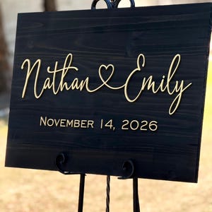 May include: A black wooden wedding sign with gold lettering. The sign reads "Nathan & Emily" with a heart symbol between the names, and "November 14, 2026" below. The sign is displayed on a black metal stand.