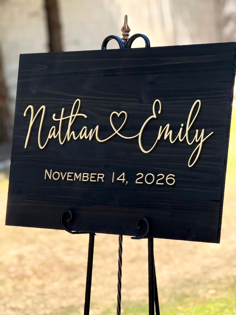 May include: A black wooden wedding sign with gold lettering. The sign reads "Nathan & Emily" with a heart symbol between the names, and "November 14, 2026" below. The sign is displayed on a black metal stand.