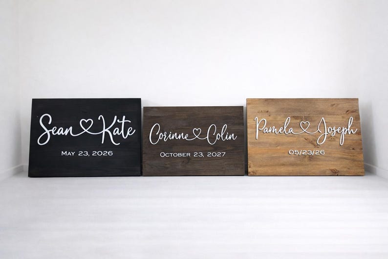 May include: Three wooden signs with names and dates. The signs are black, brown, and light brown. The names are in white script font with a heart symbol. The dates are in a simple font.