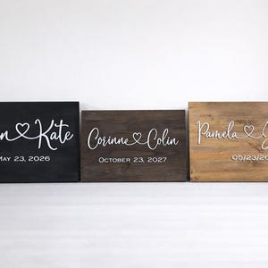 May include: Three wooden signs with names and dates. The signs are black, brown, and light brown. The names are in white script font with a heart symbol. The dates are in a simple font.