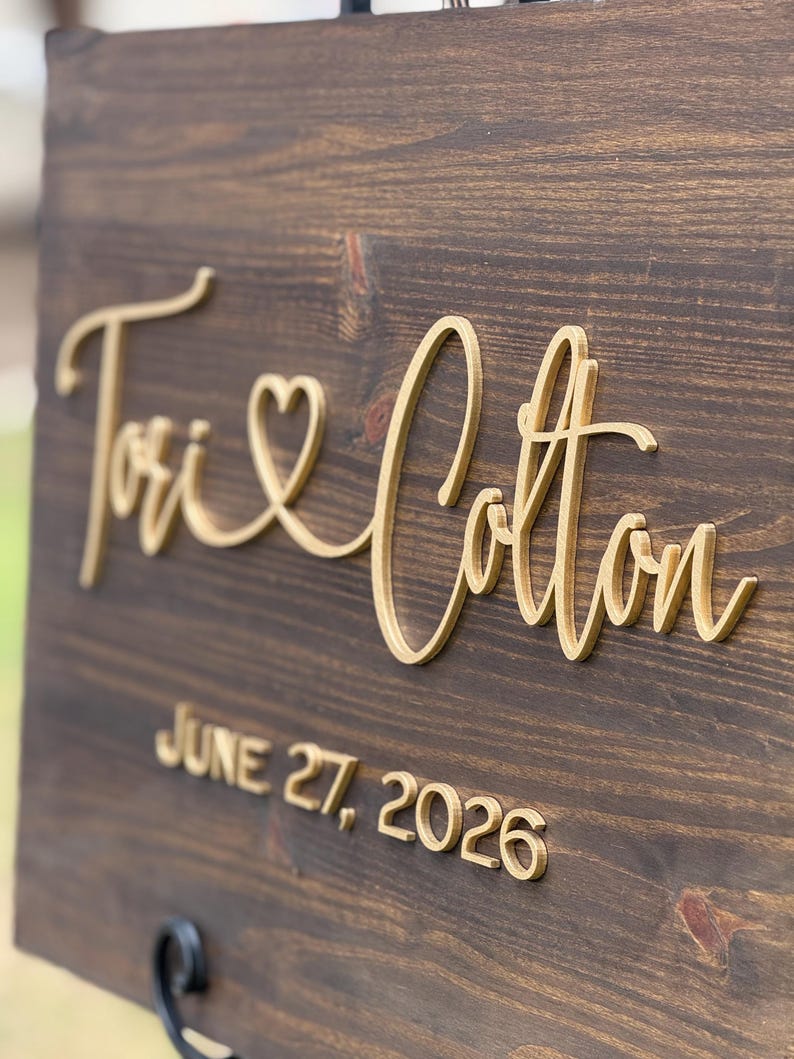 May include: A wooden sign with the names "Tori & Cotton" in elegant gold script, with a heart symbol between the names. The date "JUNE 27, 2026" is displayed below. The sign is mounted on a dark brown wooden board.