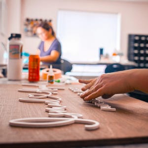 May include: A person's hands are arranging white wooden letters on a wooden board. The letters spell out a word, possibly a name or phrase. The scene takes place in a well-lit workshop with other crafting supplies visible.