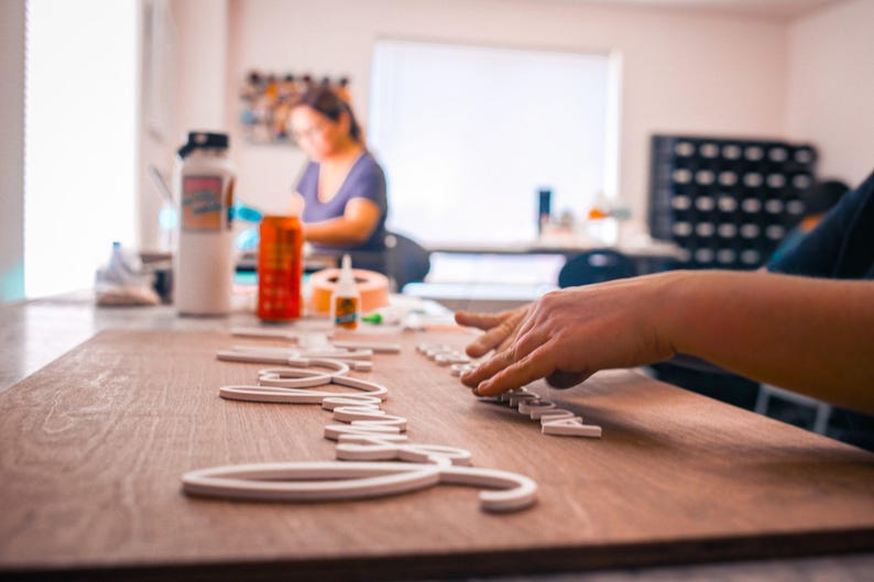 May include: A person's hands are arranging white wooden letters on a wooden board. The letters spell out a word, possibly a name or phrase. The scene takes place in a well-lit workshop with other crafting supplies visible.