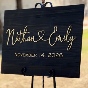 May include: A black wooden wedding sign with gold lettering. The sign reads "Nathan & Emily" with a heart symbol between the names, and "November 14, 2026" below. The sign is displayed on a decorative black metal stand.