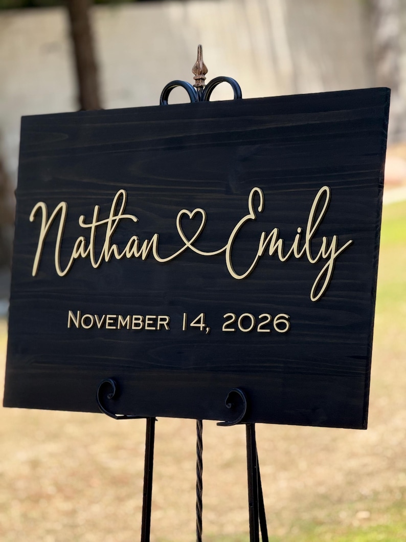 May include: A black wooden wedding sign with gold lettering. The sign reads "Nathan & Emily" with a heart symbol between the names, and "November 14, 2026" below. The sign is displayed on a decorative black metal stand.