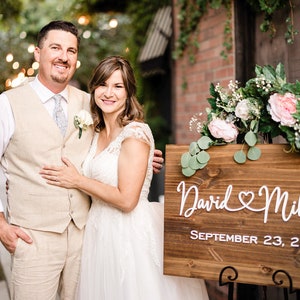 May include: A couple poses in front of a wooden sign that reads "David & Mihaela" and "September 23, 2023". The sign is decorated with flowers. The man wears a beige vest and the woman wears a white wedding dress.
