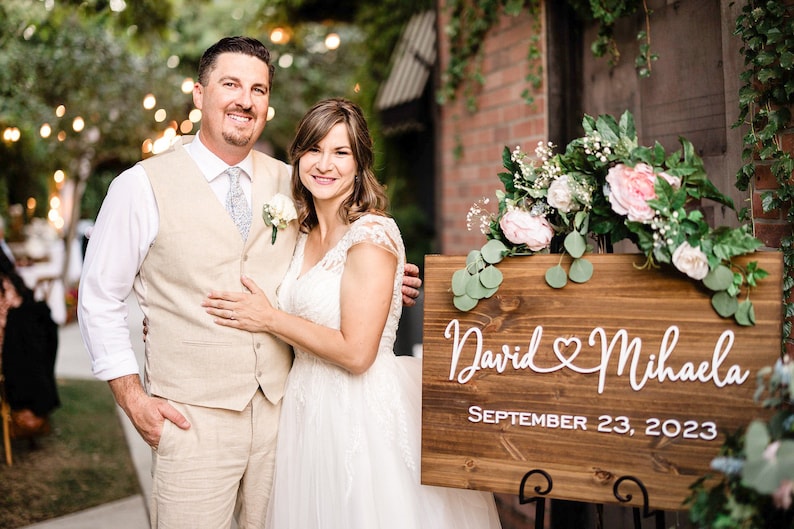 May include: A couple poses in front of a wooden sign that reads "David & Mihaela" and "September 23, 2023". The sign is decorated with flowers. The man wears a beige vest and the woman wears a white wedding dress.