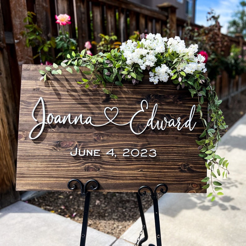 Dark Wooden sign with a couples’s 2 first names connected with a heart in white 3D lettering and wedding date.