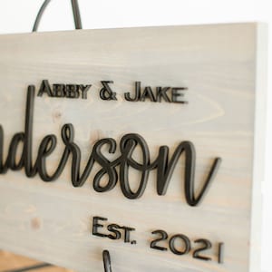 May include: A gray wooden sign with black 3D lettering that reads "Abby & Jake Anderson Est. 2021". The sign is on a black metal stand.
