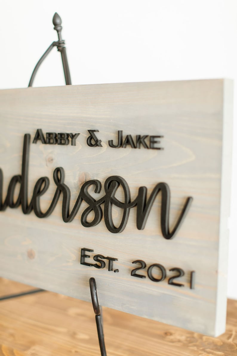 May include: A gray wooden sign with black 3D lettering that reads "Abby & Jake Anderson Est. 2021". The sign is on a black metal stand.