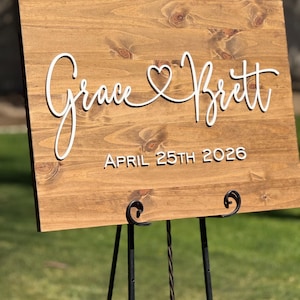 May include: A wooden sign with the names "Grace & Brett" in white script, connected by a heart symbol. The date "APRIL 25TH 2026" is below the names. The sign is displayed on a black metal easel, set against a blurred green background.