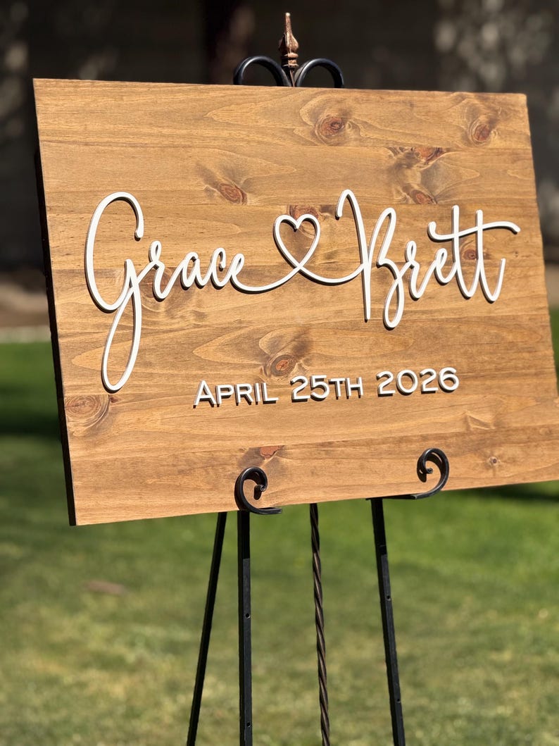 May include: A wooden sign with the names "Grace & Brett" in white script, connected by a heart symbol. The date "APRIL 25TH 2026" is below the names. The sign is displayed on a black metal easel, set against a blurred green background.
