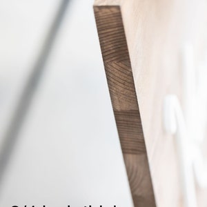 May include: Close-up of a wooden sign with a natural wood grain. The sign is 3/4 inch thick solid wood. The edge of the sign is visible, showing the layered construction of the wood. The background is blurred, with some greenery visible.
