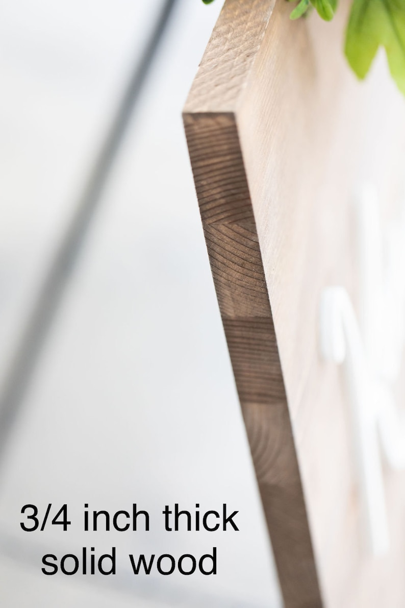 May include: Close-up of a wooden sign with a natural wood grain. The sign is 3/4 inch thick solid wood. The edge of the sign is visible, showing the layered construction of the wood. The background is blurred, with some greenery visible.