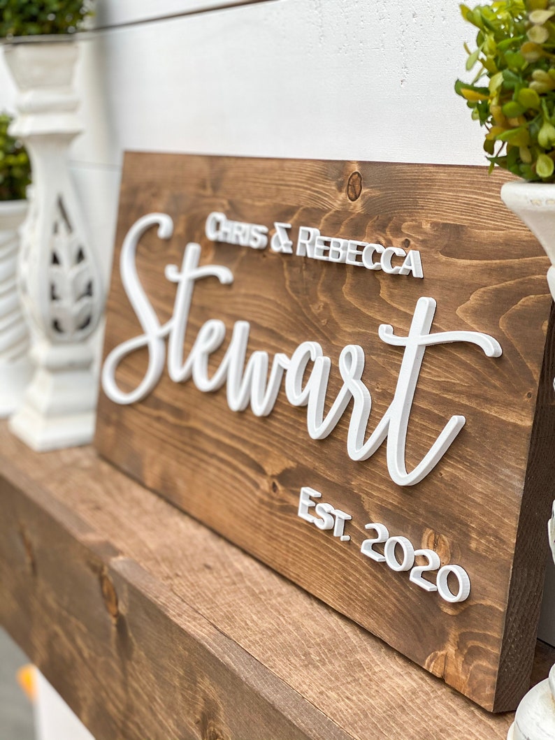 May include: A brown wooden sign with white raised lettering that reads "Chris & Rebecca Stewart Est. 2020".