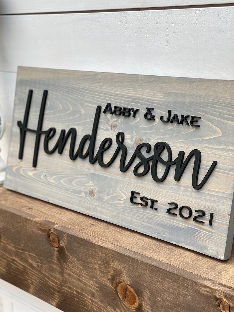 May include: A gray wooden sign with the text "Henderson" in black raised letters. The text "Abby & Jake" and "Est. 2021" are also in black raised letters.