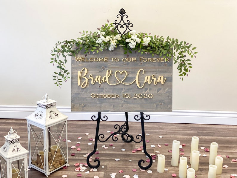 Custom wood wedding sign with gray stain and gold 3D letters. Contains Welcome to our Wedding or Welcome to our Forever with couple's names connected with a heart and wedding date below.