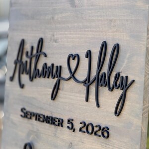 May include: A wooden sign with the names "Anthony & Haley" in black script, joined by a heart symbol. Below, the date "SEPTEMBER 5, 2026" is displayed in a bold, sans-serif font. The sign has a gray-toned finish.