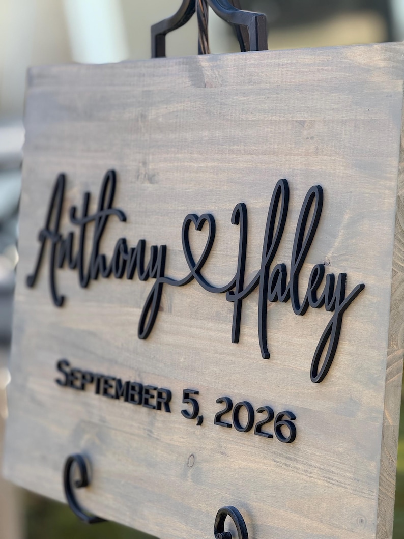May include: A wooden sign with the names "Anthony & Haley" in black script, joined by a heart symbol. Below, the date "SEPTEMBER 5, 2026" is displayed in a bold, sans-serif font. The sign has a gray-toned finish.