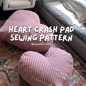 May include: Two large heart-shaped floor cushions featuring a pink and white striped fabric pattern. The text HEART CRASH PAD SEWING PATTERN and @HANNAHADELESHOME is overlaid on the image. The cushions are soft, stuffed, and rounded.