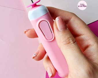 May include: A pink handheld tool with a white top and a small needle-like point. The tool has a button on the side and is held in a hand with white flowers on the fingernails.