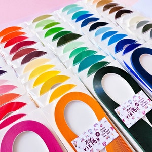 Puede incluir: Surtido de tiras de papel para quilling de colores en varios tonos, dispuestas en filas sobre un fondo rosa. Cada tira tiene forma de semic&iacute;rculo y est&aacute; etiquetada con el nombre de la marca "Tilly Viktor".