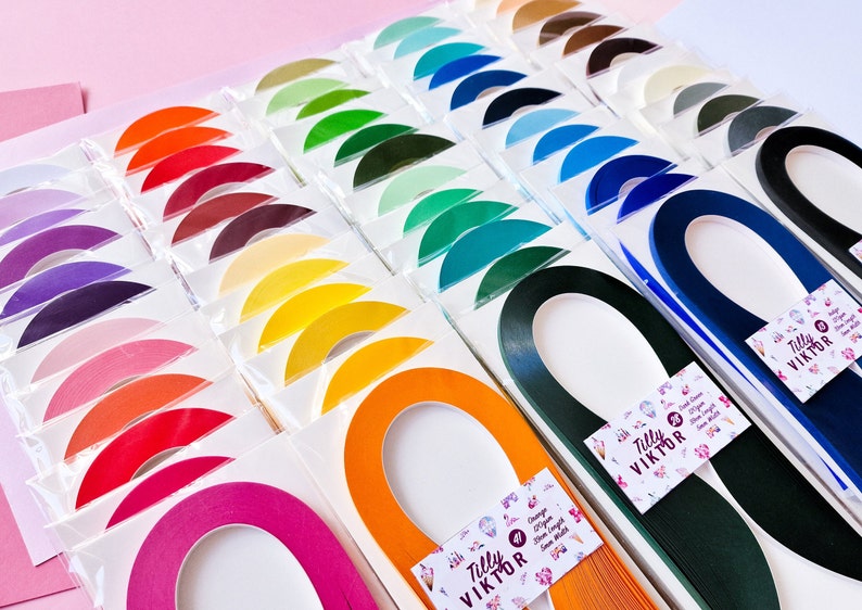 Puede incluir: Surtido de tiras de papel para quilling de colores en varios tonos, dispuestas en filas sobre un fondo rosa. Cada tira tiene forma de semic&iacute;rculo y est&aacute; etiquetada con el nombre de la marca "Tilly Viktor".