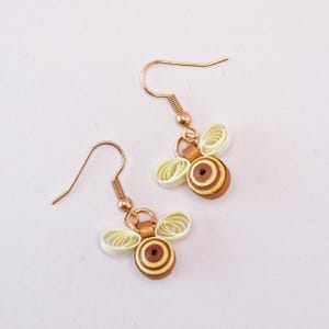 May include: A pair of handmade bee earrings. The earrings feature a gold-colored hook and a bee design made of rolled paper in shades of brown, yellow, and white. The bee's wings are made of white paper.