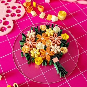Peut inclure: Un bouquet de quilling en papier fait à la main avec des fleurs jaunes, orange et blanches et des feuilles vertes. Le bouquet est disposé sur une base circulaire transparente, avec une tige en papier vert. Le fond est une grille rose.