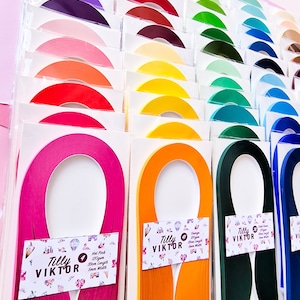 Puede incluir: Surtido de tiras de papel de colores para manualidades, dispuestas en filas sobre un fondo rosa. Cada tira est&aacute; etiquetada con el nombre de la marca "Tilly Viktor" y un n&uacute;mero. Las tiras son de varios colores, incluyendo rosa intenso, naranja, verde y azul.