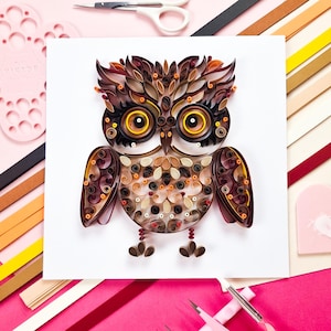 Puede incluir: Una detallada obra de arte de búho con quilling de papel. El búho presenta tiras de papel marrón, beige y naranja, con grandes ojos amarillos. La obra de arte está sobre un fondo blanco, con materiales de manualidades.
