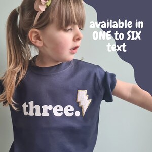 May include: A navy blue t-shirt with a white lightning bolt and the word "three." The shirt is available in sizes one to six.
