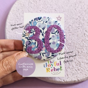 May include: A purple glitter number 30 on a white fabric background with a floral pattern. The fabric is a Liberty print. The text "Pick your glitter text" and "Pick your Liberty print fabric" is written on the card. The text "The Joyful Rebel" is written on the card in colourful letters.